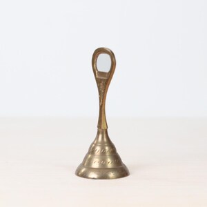 May include: A vintage brass bell with a teardrop-shaped handle and a flared base. The bell has a detailed, textured design around the base and a smooth, polished finish. The bell is likely used for calling or signaling.