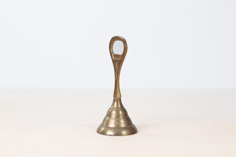 May include: A vintage brass bell with a teardrop-shaped handle and a flared base. The bell has a detailed, textured design around the base and a smooth, polished finish. The bell is likely used for calling or signaling.