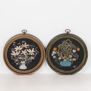 May include: Two circular wall art pieces featuring embroidered floral arrangements on black fabric. The left piece has a light-colored vase with pink and white flowers. The right piece has a blue vase with orange and green foliage, signed NAK 72.