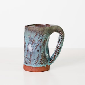 Może przedstawiać: Ręcznie robiony ceramiczny kubek o unikalnym designie. Kubek ma teksturowane, turkusowe i brązowe szkliwo z czerwoną podstawą i wzorzystym uchwytem. Kubek ma około 10 cm wysokości.