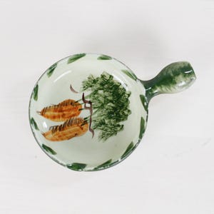 May include: A small, round ceramic dish with a single handle, featuring a hand-painted design of two orange carrots with green leafy tops on a cream-colored background, accented by green leaf motifs around the interior rim of the bowl.
