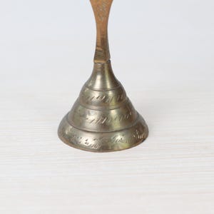 May include: A vintage brass bell with a conical base and a slender stem. The bell has etched detailing around the base and a warm, aged patina. The bell is likely used for decorative purposes.