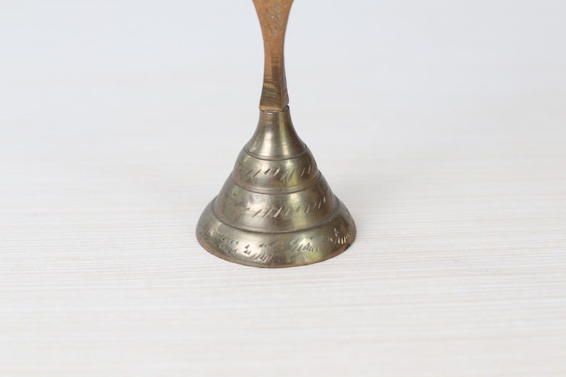May include: A vintage brass bell with a conical base and a slender stem. The bell has etched detailing around the base and a warm, aged patina. The bell is likely used for decorative purposes.