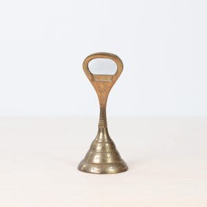 May include: A vintage brass bottle opener with a distinctive design. The opener features a loop handle and a conical base with etched detailing. The brass has a warm, aged patina, suggesting its vintage origin. The opener stands upright on a light-colored surface.