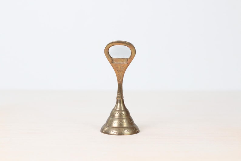 May include: A vintage brass bottle opener with a distinctive design. The opener features a loop handle and a conical base with etched detailing. The brass has a warm, aged patina, suggesting its vintage origin. The opener stands upright on a light-colored surface.