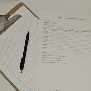 May include: A clipboard with lined paper, a black pen, and a printed document titled "Estrogen Patch Symptom Tracker (Weekly)". The document includes sections for tracking mood, hot flashes, sleep, and energy levels.