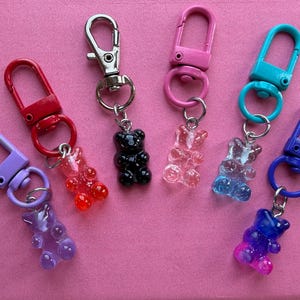May include: Six keychains featuring gummy bear charms in various colours including purple, red, black, clear, light blue, and a blue-to-pink gradient. Each charm is attached to a matching coloured lobster clasp by a small silver metal jump ring.