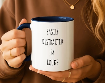 Op de afbeelding: Witte keramische mok met een marineblauwe binnenkant en handvat. De mok heeft de zwarte tekst "EASILY DISTRACTED BY ROCKS". De persoon draagt een bruine trui. De mok is een standaard formaat.