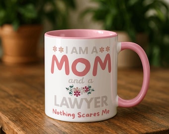 May include: White ceramic mug with a pink interior and handle. The mug features the text "I AM A MOM and a LAWYER Nothing Scares Me" in pink and white, with floral accents. The mug is on a wooden surface.