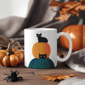 May include: White ceramic mug with a Halloween-themed design. The design features a black cat on an orange pumpkin, with another black cat on a teal base. A small pumpkin and a toy spider are on the wooden surface.