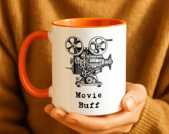 May include: White ceramic mug with an orange interior and handle. The mug features a black and white illustration of a vintage movie camera with the words "Movie Buff" printed below the image. The mug is held in a person's hands.