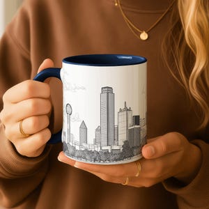 May include: White ceramic mug with a navy blue interior and handle. The mug features a black and white line drawing of a city skyline. The mug is held by a person.