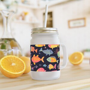 Op de afbeelding: Witte mason jar met een handvat en een zilveren deksel, versierd met een kleurrijk vissenpatroon op een donkerblauwe achtergrond. Inclusief een wit rietje. De pot staat op een houten oppervlak met sinaasappelschijfjes.