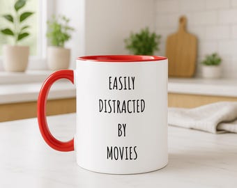 May include: White ceramic mug with a red interior and handle. The mug features the text "EASILY DISTRACTED BY MOVIES" in black, centered on the front. The mug is on a white surface, with plants and a cutting board in the background.
