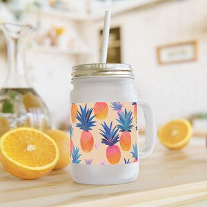 Op de afbeelding: Witte mason jar-mok met een handvat en een zilveren deksel, versierd met een kleurrijk ananaspatroon. De ananassen zijn in tinten van oranje, roze en geel met blauwe bladeren. Een wit rietje is in de mok gestoken.
