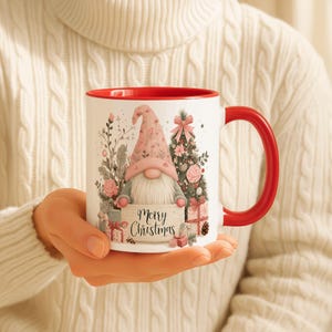 May include: White ceramic mug with a red interior and handle. The mug features a gnome design with a pink hat, holding a sign that reads "Merry Christmas". The design includes a Christmas tree, gifts, and floral elements.