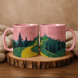 May include: Two pink ceramic mugs featuring a landscape painting of a yellow brick road winding through a green forest under a pink sky. The mugs have pink handles and are on a wooden slice.