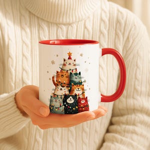 May include: White ceramic mug with a red interior and handle. The mug features a whimsical illustration of a Christmas tree made of cats, adorned with string lights and a star on top. Snowflakes are scattered around the tree.