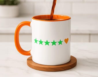 May include: White ceramic mug with an orange interior and handle, featuring green star graphics and an orange heart. The mug sits on a wooden coaster, with coffee being poured into it. A kitchen setting is in the background.