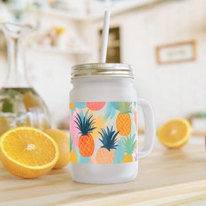 Könnte beinhalten: Weißes Einmachglas mit silbernem Deckel, Henkel und weißem Strohhalm. Das Glas ist mit einem farbenfrohen Ananasmuster in Orange, Pink und Türkis verziert. Perfekt für kalte Getränke, Smoothies oder Eistee.