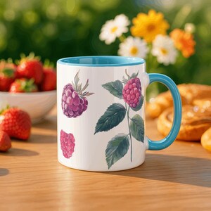 Peut inclure: Mug en céramique blanche avec intérieur et anse bleus, orné d'illustrations à l'aquarelle de framboises et de feuilles vertes. Le mug est posé sur une surface en bois, avec des fraises et des fleurs en arrière-plan.