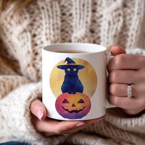 May include: White ceramic mug featuring a black cat wearing a witch hat, sitting atop a jack-o'-lantern. The design includes a full moon backdrop. The mug is held by a person wearing a cream-colored sweater.