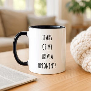 May include: White ceramic mug with a black interior and handle. The mug features the text "TEARS OF MY TRIVIA OPPONENTS" in black, centered on the front. The mug is on a wooden surface with a newspaper and a cream-coloured knitted item in the background.