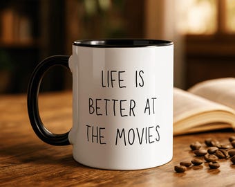 May include: White ceramic mug with a black interior and handle. The mug features the text "LIFE IS BETTER AT THE MOVIES" in black lettering. Coffee beans are scattered on the wooden surface next to the mug.