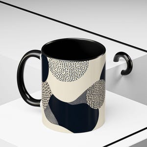 May include: A ceramic coffee mug with a black handle and interior. The exterior features a cream-colored background with navy blue and dotted black abstract shapes. The mug is displayed on a white surface.