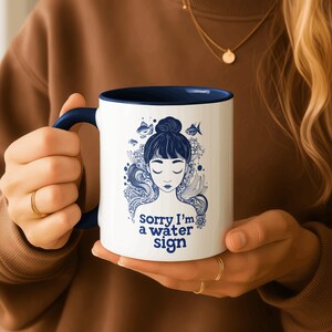 May include: White ceramic mug with a navy blue interior and handle. The mug features a blue illustration of a woman with closed eyes, fish, and the text "Sorry I'm a water sign."