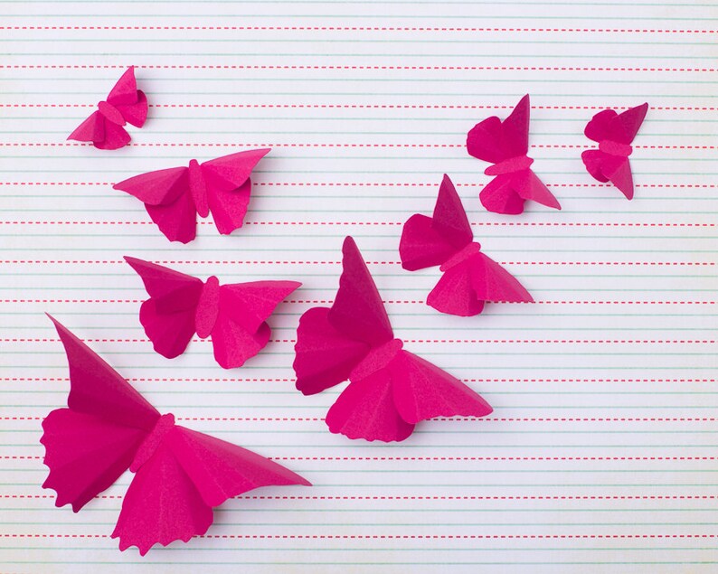 May include: A collection of pink paper butterflies arranged on a lined sheet of paper. The butterflies are all different sizes and shapes, and they are all facing different directions.