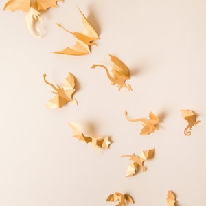 May include: A collection of ten paper dragon cutouts in various sizes and poses, all in a light gold colour, against a light beige background.