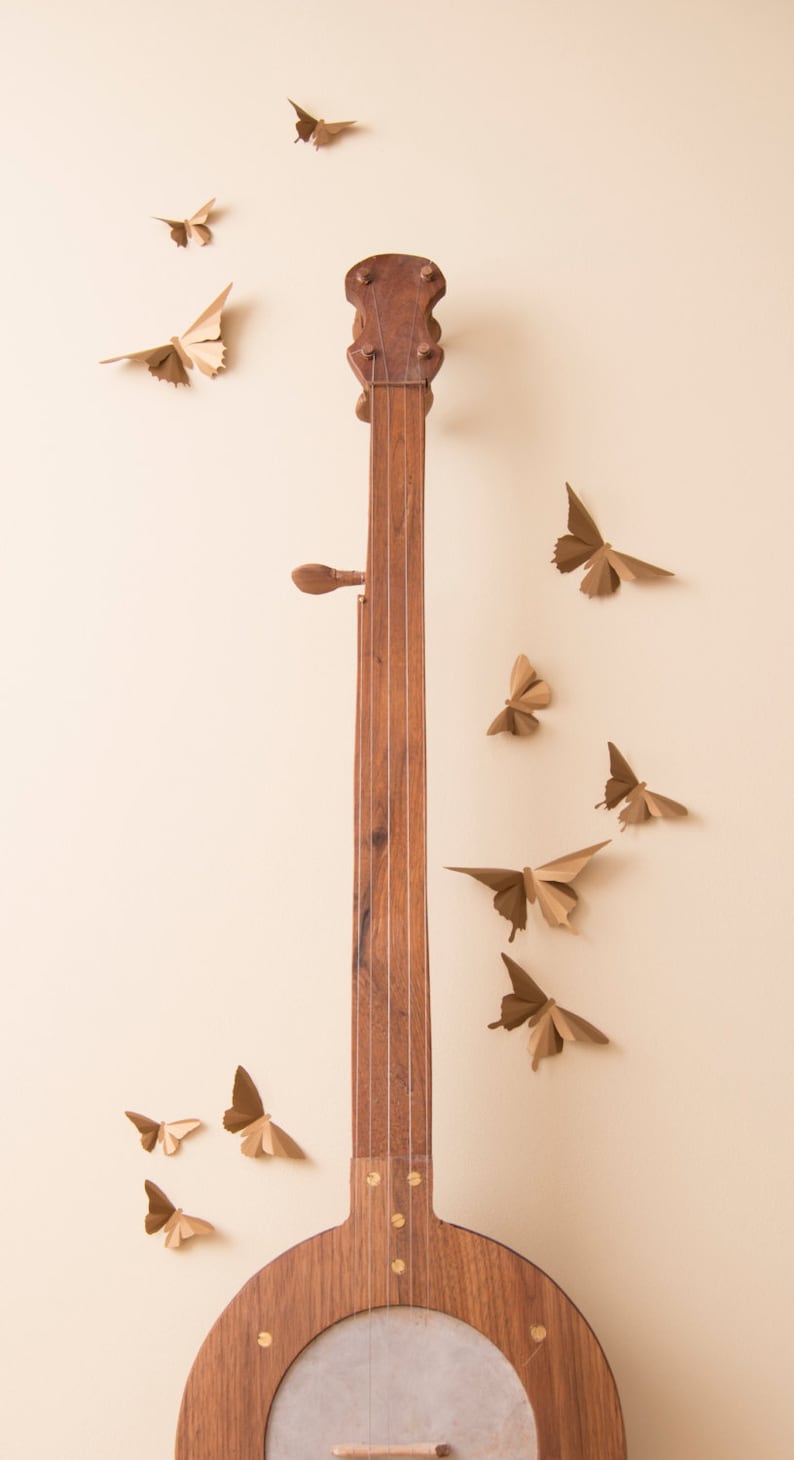 May include: A wooden banjo with a light brown finish and gold accents. The banjo is surrounded by paper butterflies in various shades of brown.