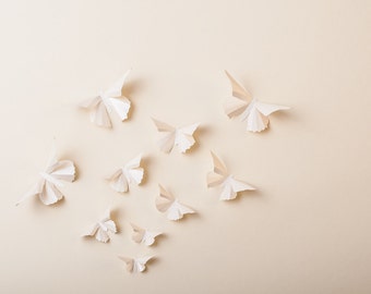 May include: A collection of white paper butterflies arranged on a beige background. The butterflies are all different sizes and shapes, and they are all facing different directions.
