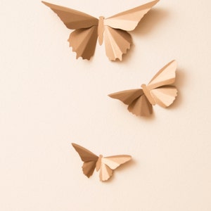 May include: Three paper butterflies in various sizes, all in a light brown color, are arranged on a light beige background.