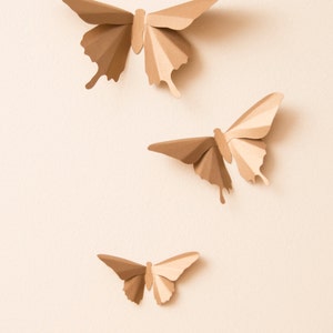 May include: Three paper butterflies in various sizes, all in a light brown color, are arranged on a light beige background.