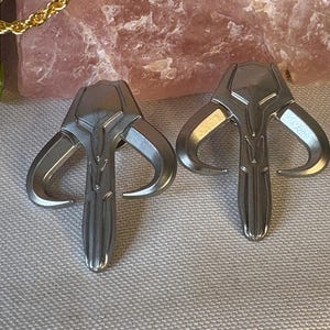 May include: Two silver-colored Mandalorian helmet pendants. Each pendant features a stylized, angular design with curved horns and a central vertical element. The pendants are displayed on a light gray surface, with a gold chain and a pink stone in the background.