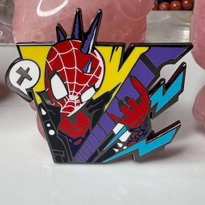 May include: A colorful enamel pin featuring a stylized Spider-Man design. The pin showcases a red and white spiderweb mask, a black jacket, and a yellow and purple background. The design includes a speech bubble with a white X.