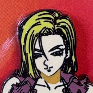 Puede incluir: Primer plano de un pin que representa a un personaje femenino de anime con cabello rubio y una chaqueta morada. El personaje tiene una expresión seria sobre un fondo rojo y naranja. El pin está delineado en negro.