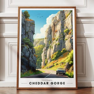 May include: A framed print of Cheddar Gorge, UK. The artwork shows a road through a dramatic gorge with high rock formations. A vintage car is on the road. The sky is blue with birds. The print is in a wooden frame.