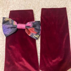 Może przedstawiać: Dwa bordowe aksamitne mankiety do butów z kwiatową kokardą. Kokarda ma mieszankę kolorów, w tym fioletowy, różowy i czerwony. Mankiety są prostokątne i mają miękką, pluszową fakturę.