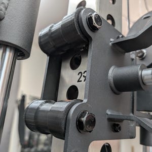 Può includere: Primo piano di un componente di una macchina per sollevamento pesi. L'immagine mostra un rullo nero e cilindrico attaccato a un telaio metallico grigio con bulloni neri. Il numero "29" è visibile sul telaio. Lo sfondo è sfocato, suggerendo un ambiente di palestra.