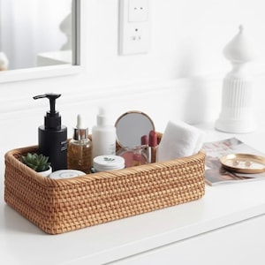 Peut inclure: Un panier rectangulaire en rotin tressé rempli de produits de beauté, dont un flacon pompe noir, un petit miroir et une plante succulente. Le panier est posé sur une surface blanche, suggérant une salle de bain ou une coiffeuse.