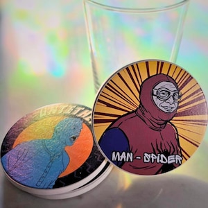 May include: Two round coasters with cartoon illustrations and a clear glass. One coaster features a blue figure with the text "UNTITLED". The other coaster has a character in a red hood with the text "MAN-SPIDER".
