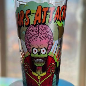 May include: A clear glass featuring a colorful graphic of a Martian alien with a large brain and the words "MARS ATTACKS" in orange. The alien is depicted in a red and green outfit with a skeletal face. The glass is cylindrical.