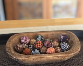 Op de afbeelding: Een kleine, ovale, met de hand gesneden houten deegkom gevuld met een verzameling ronde, met stof beklede bollen. De bollen zijn voorzien van diverse ruit- en blokpatronen in tinten oranje, bruin, blauw, wit en paars.