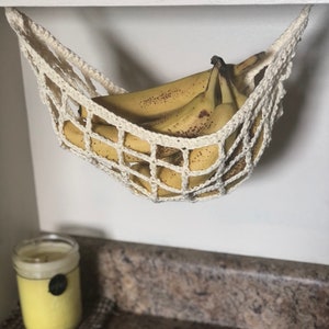 May include: A white crocheted hammock hanging from a white cabinet, holding a bunch of yellow bananas.