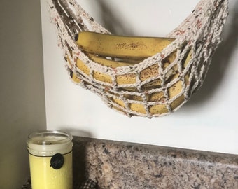 Op de afbeelding: Een witte gehaakte hangmat die aan een wit rek hangt, met een tros gele bananen erin. Een gele kaars in een glazen pot staat op een bruin aanrecht.
