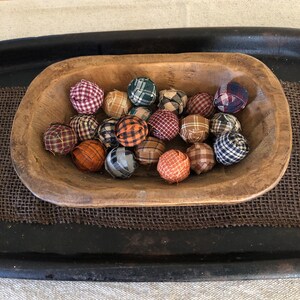 Pu&ograve; includere: Una collezione di piccole sfere di tessuto rotonde con vari motivi a quadri e scozzesi nei toni del rosso, blu, arancione, marrone e verde, tutte contenute all'interno di una ciotola di legno allungata, rustica e naturale con interno intagliato.