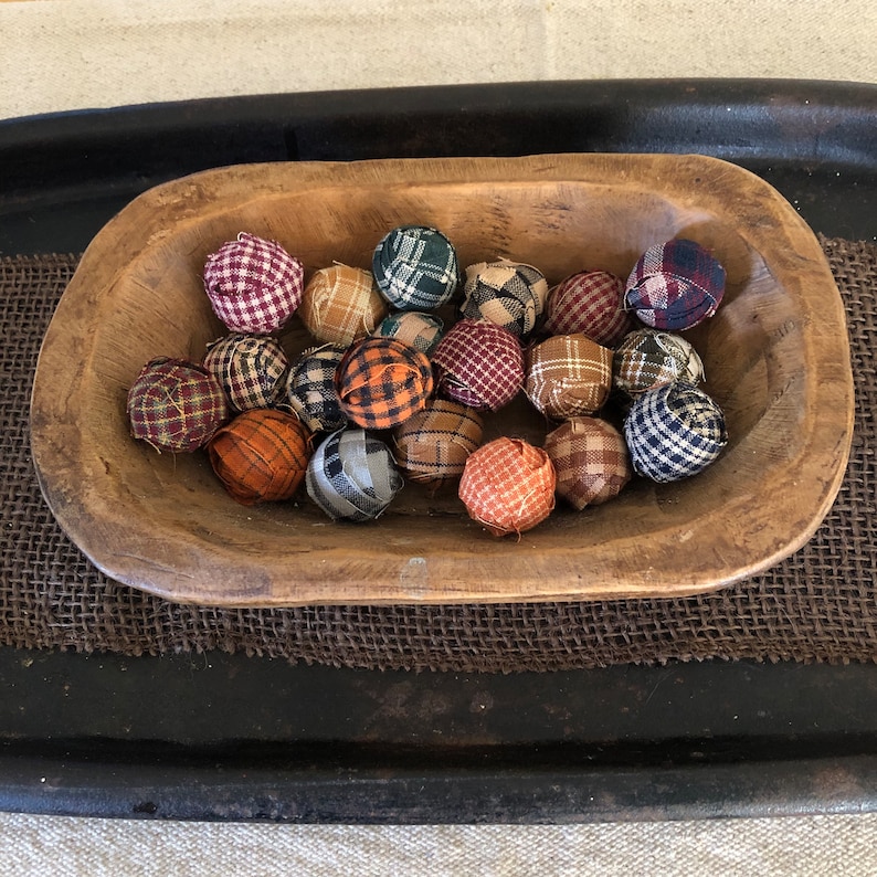 Pu&ograve; includere: Una collezione di piccole sfere di tessuto rotonde con vari motivi a quadri e scozzesi nei toni del rosso, blu, arancione, marrone e verde, tutte contenute all'interno di una ciotola di legno allungata, rustica e naturale con interno intagliato.